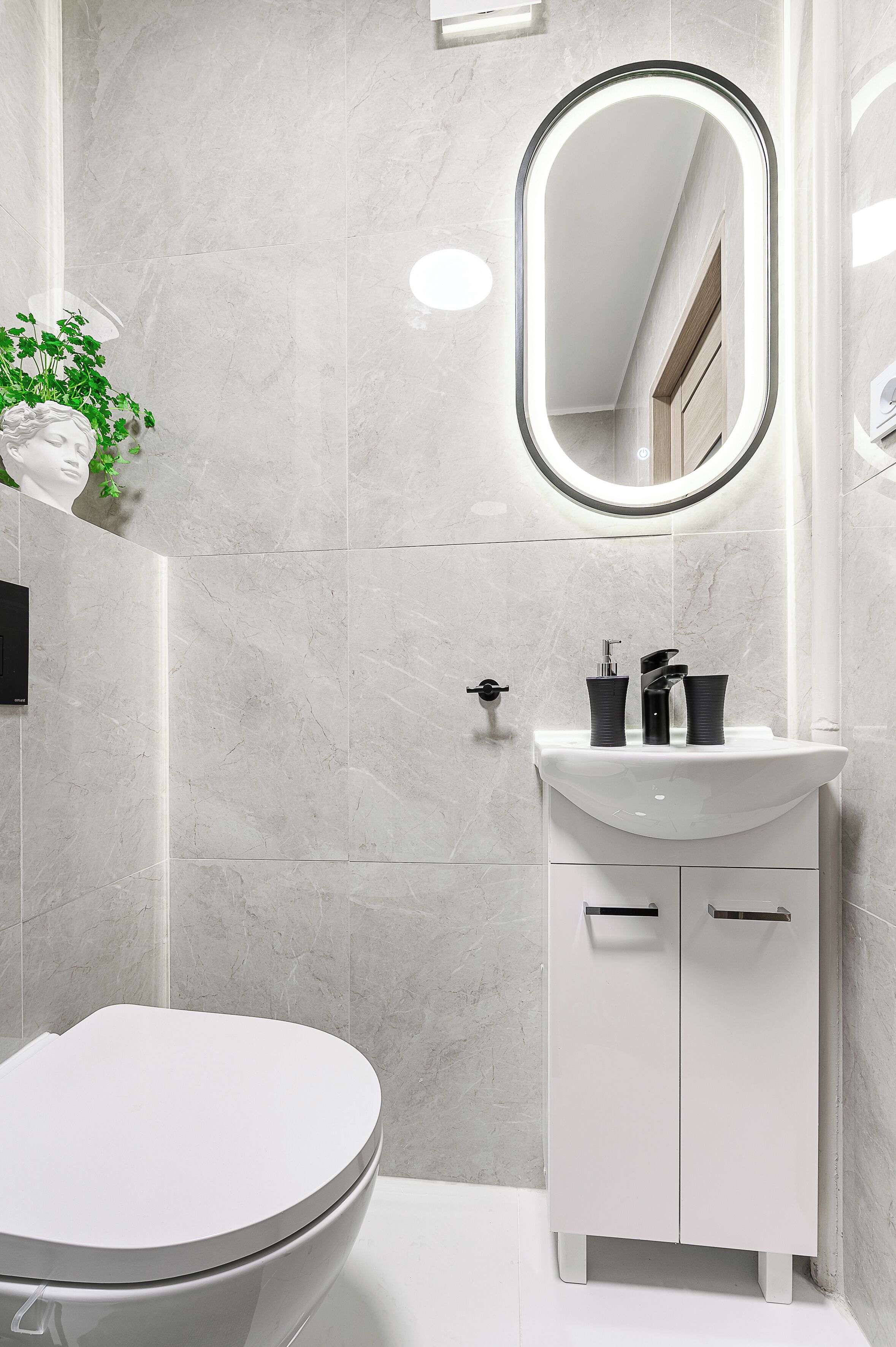 A modern bathroom with a white toilet, a white sink with black fixtures, and a mirror with a light.
