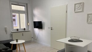 Een moderne, minimalistische kamer met een witte deur, een flatscreen televisie aan de muur, een klein rond tafeltje met een metalen schaal, en een grijze bank.