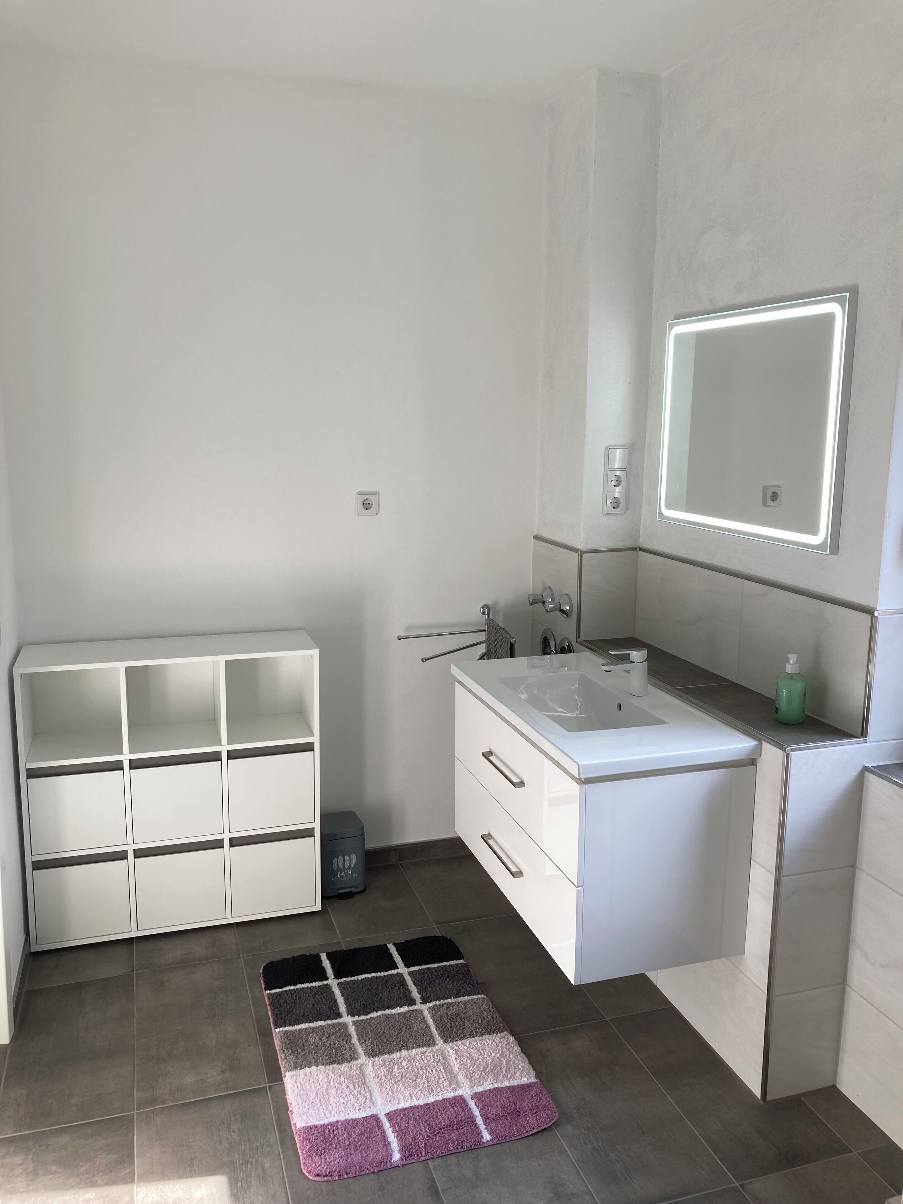 A modern bathroom with a white vanity, a large mirror with LED lighting, a built-in shelf, and a patterned rug on the floor.