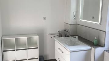 A modern bathroom with a white vanity, a large mirror with LED lighting, a built-in shelf, and a patterned rug on the floor.