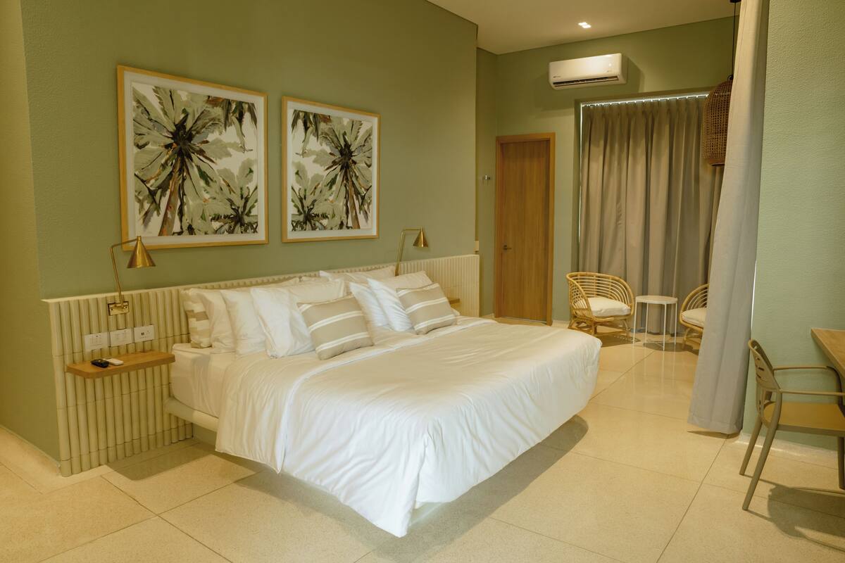 A bedroom with a large bed, two wall-mounted artworks, a bedside table with a lamp, a chair, and a small table.