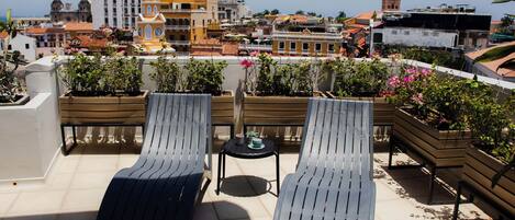A rooftop terrace with two lounge chairs and a small table, overlooking a cityscape with historic buildings and a clock tower.