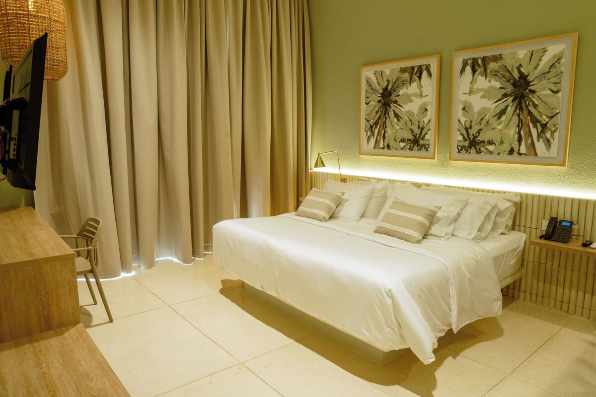 A hotel room with a large bed, a bedside table with a lamp, a chair, and two framed artworks on the wall.
