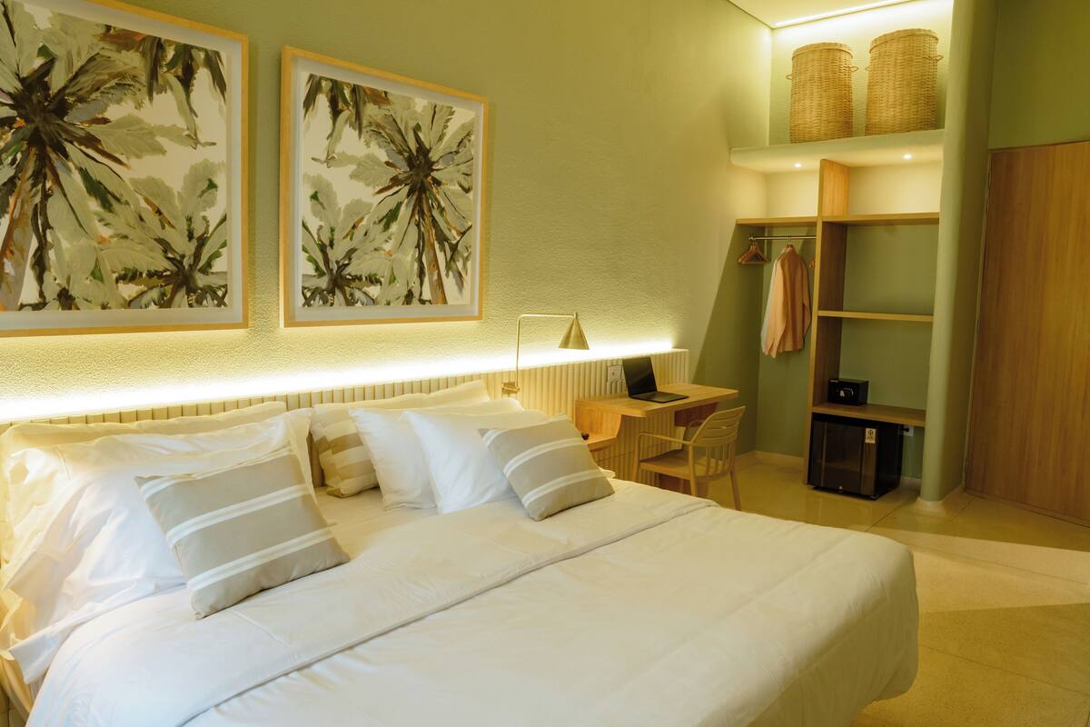 A neatly arranged hotel room with a large bed, a desk, and a chair. There are two framed artworks on the wall, a lamp, and a wardrobe.