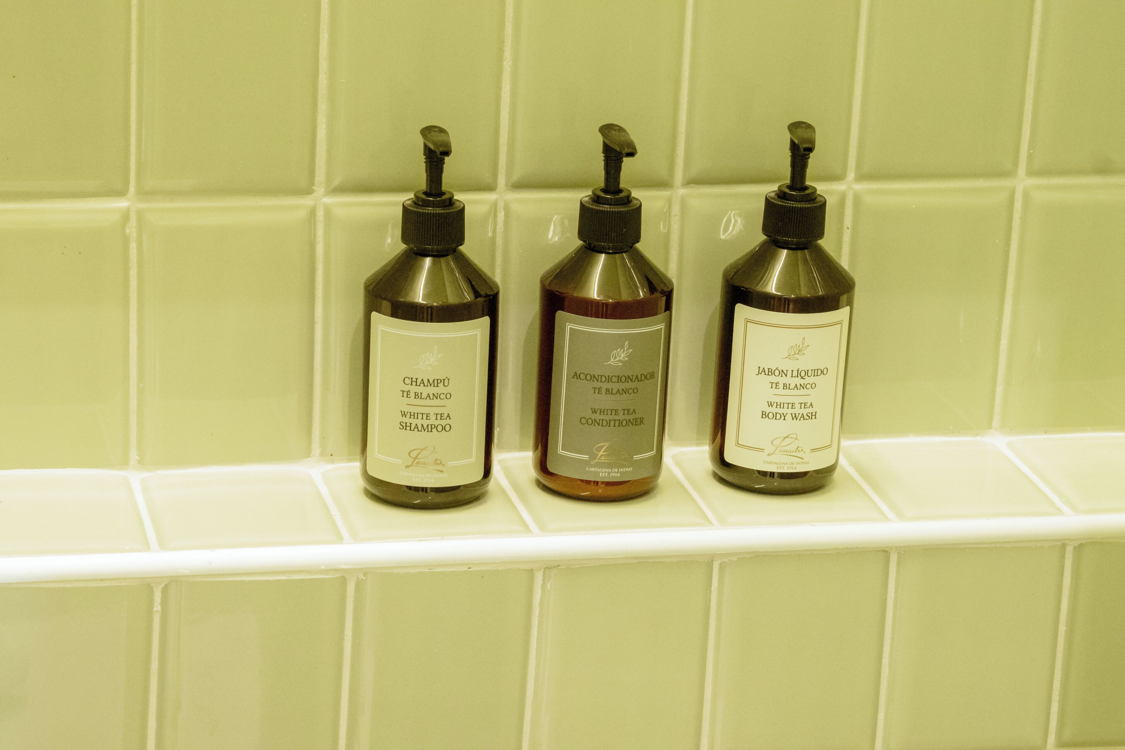 Three bottles of white tea body wash on a white shelf against a tiled wall.
