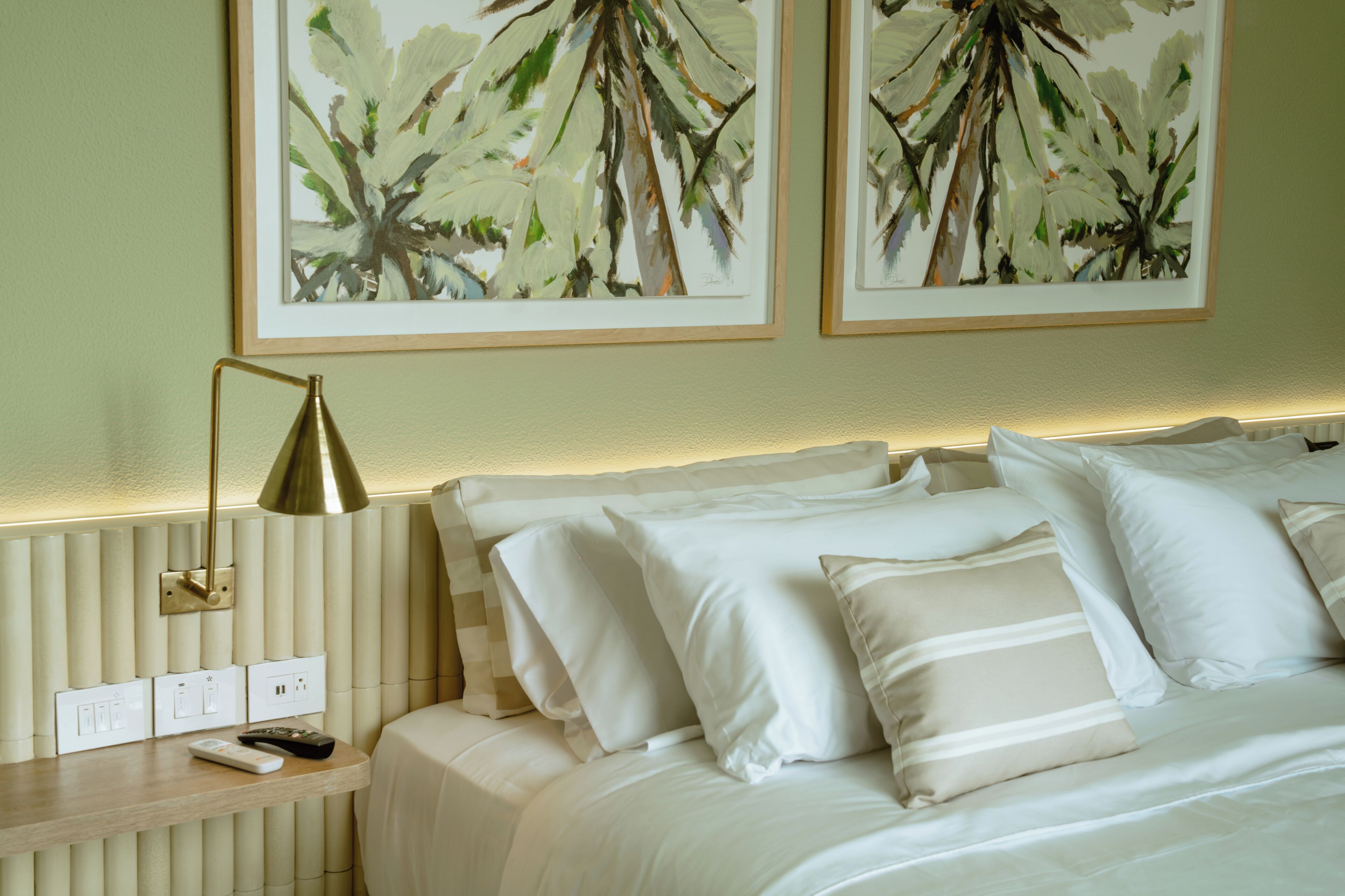 A hotel room with a bed, pillows, a bedside table with a lamp, and two framed paintings on the wall.