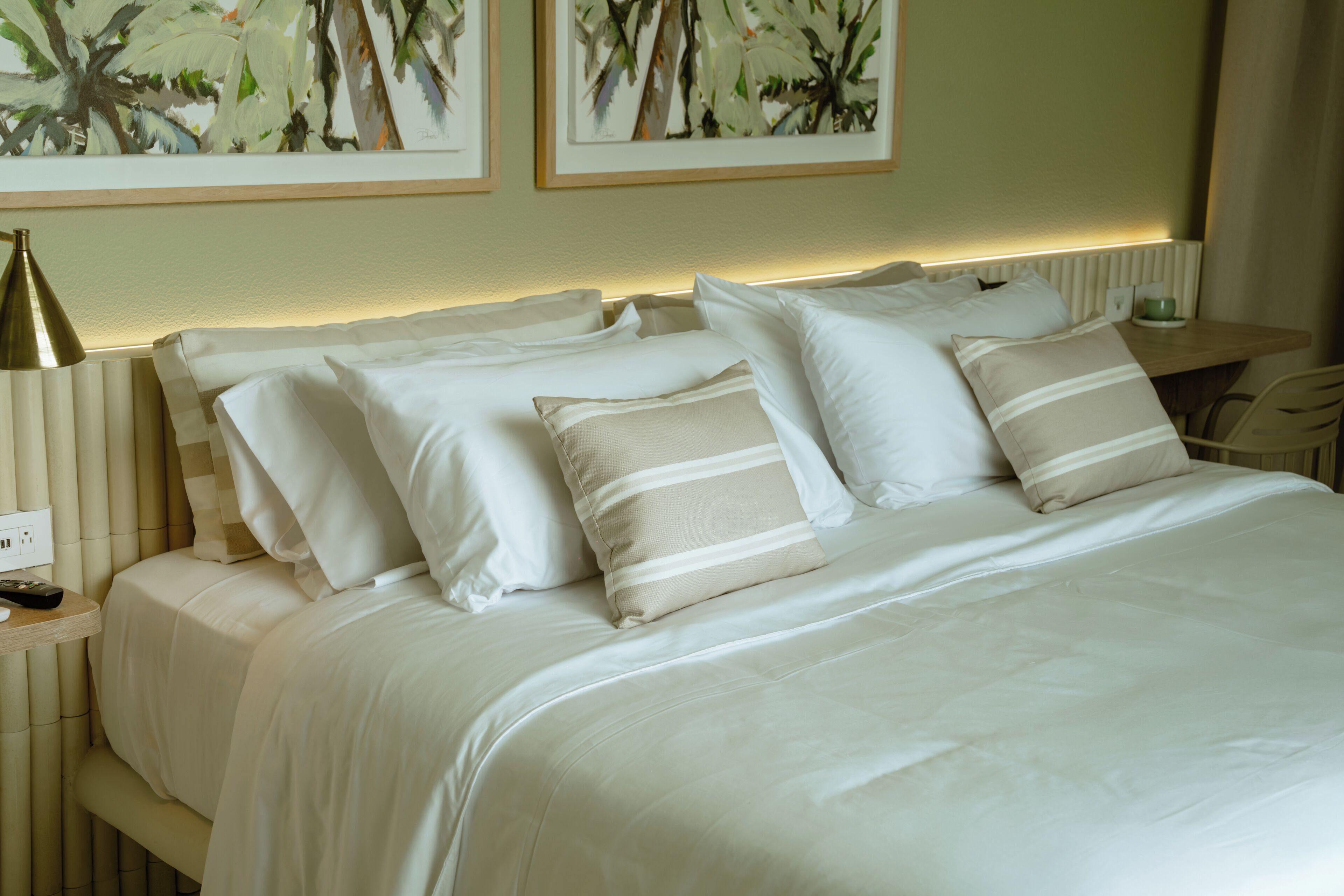 A neatly made bed with white linens and striped pillows, a bedside lamp, and a small table with a cup.