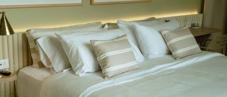 A neatly made bed with white linens and striped pillows, a bedside lamp, and a small table with a cup.