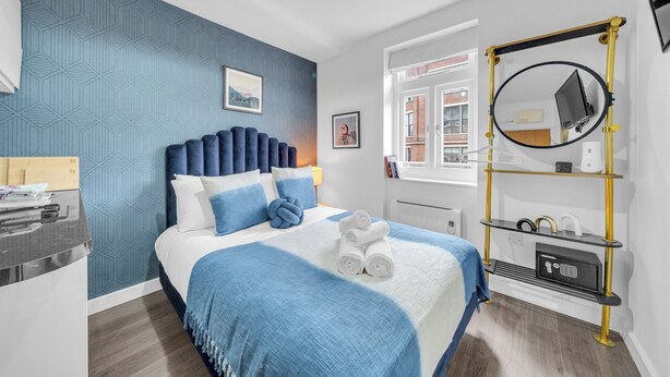 Quarto de hotel moderno com parede azul estampada em padrão geométrico, cama com roupa de cama azul, mesa de cabeceira com abajur e espelho com moldura dourada.