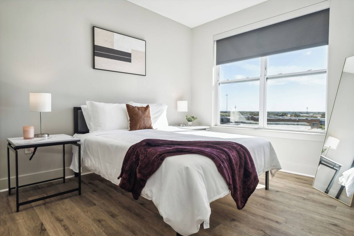 Une chambre d’hôtel moderne avec une grande fenêtre, un lit soigneusement fait, une table de chevet avec une lampe et une œuvre d’art encadrée au mur.