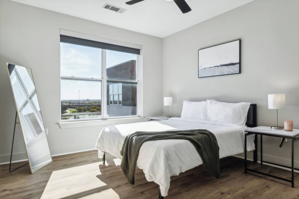 Une chambre moderne avec une grande fenêtre, un lit, des tables de chevet et une vue sur le paysage urbain.