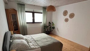 A bedroom with a bed, a wooden floor, a window with curtains, a bedside table with a lamp, and a potted plant. - Quiet apartment for commuters & nature lovers - parking lot (Duisburg)