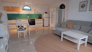 A modern kitchen with white cabinets, a green backsplash, and a built-in oven. There is a white dining table with a chair, a gray sofa, and a white coffee table in the living area. - Quiet apartment for commuters & nature lovers - parking lot (Duisburg)