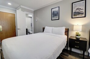 Un lit bien fait, avec du linge de lit blanc, une table de chevet avec une lampe et une petite plante, et deux tableaux encadrés accrochés au mur.