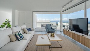 A modern living room with a white sofa, a coffee table, and a TV. There is a balcony with a view of the sky and clouds. - Villa Cala - Casangular by Upper (Adeje)