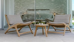 Deux fauteuils en osier avec des coussins gris, une petite table en bois et une porte en verre qui reflète un lit et de la verdure.