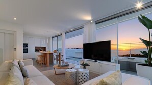 Un soggiorno moderno con una grande TV, posti a sedere comodi, un'area pranzo e vista sul tramonto attraverso ampie finestre.