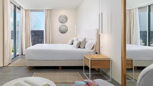 Una camera d'albergo moderna con un letto grande, un comodino, un angolo lettura con un libro e una coperta, e vista sull'esterno grazie a ampie finestre.