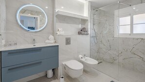 Une salle de bain moderne avec un sol en marbre, un lavabo blanc avec un meuble bleu, des toilettes, un bidet et une douche à parois de verre.