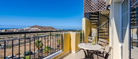 A balcony with a table and chairs, a bottle of wine, and a view of the sea and distant buildings.