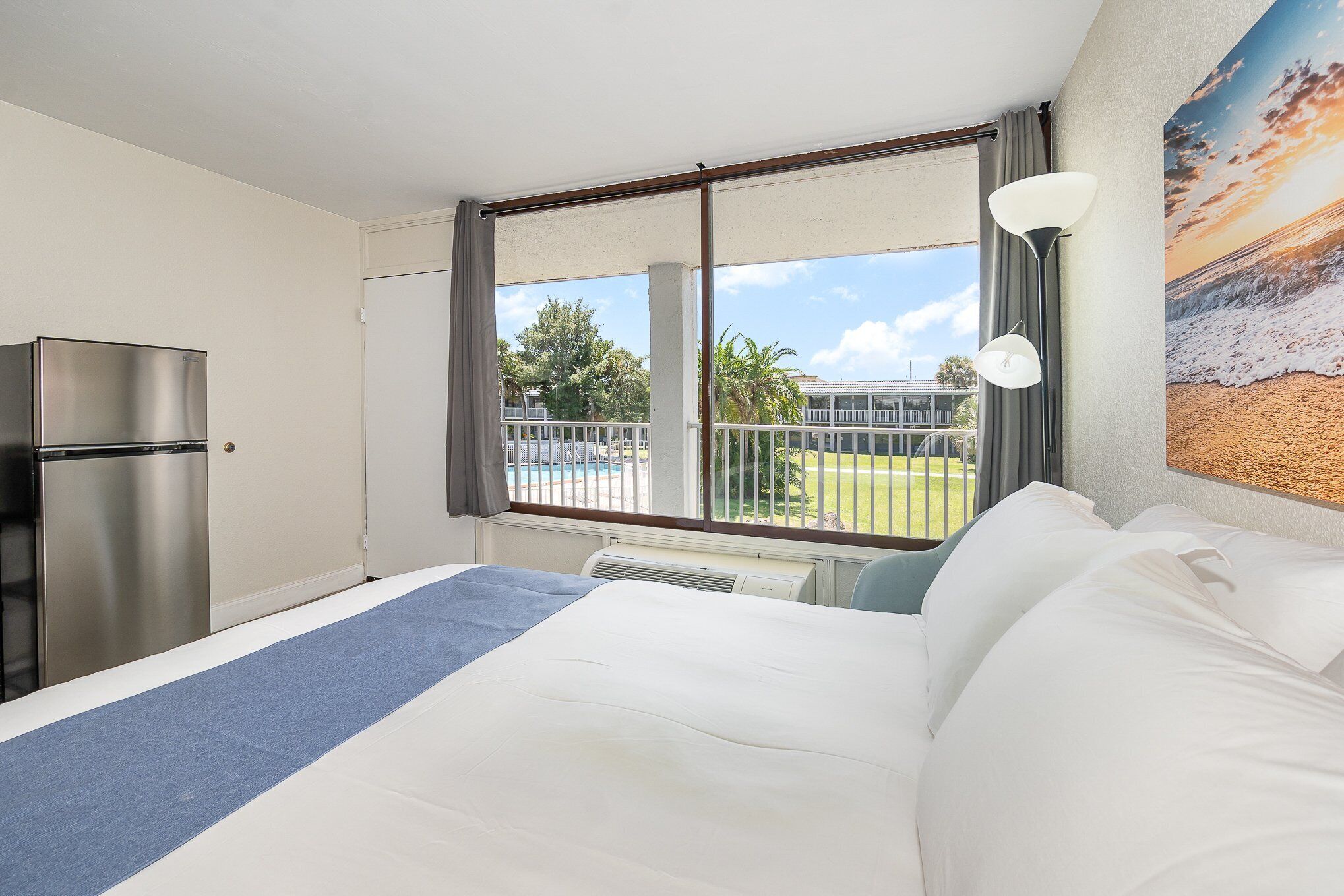 Hotelkamer met een bed, een koelkast, een raam met uitzicht op een zwembad en groen, en een schilderij aan de muur.