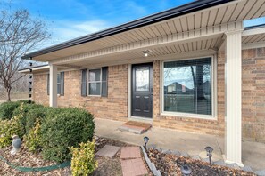 A two-story brick house with a covered porch, a front door with a doormat, and a small garden area with a bird feeder. - 1-Acre Retreat, Fire Pit, Near ASU & Hospital (Jonesboro)