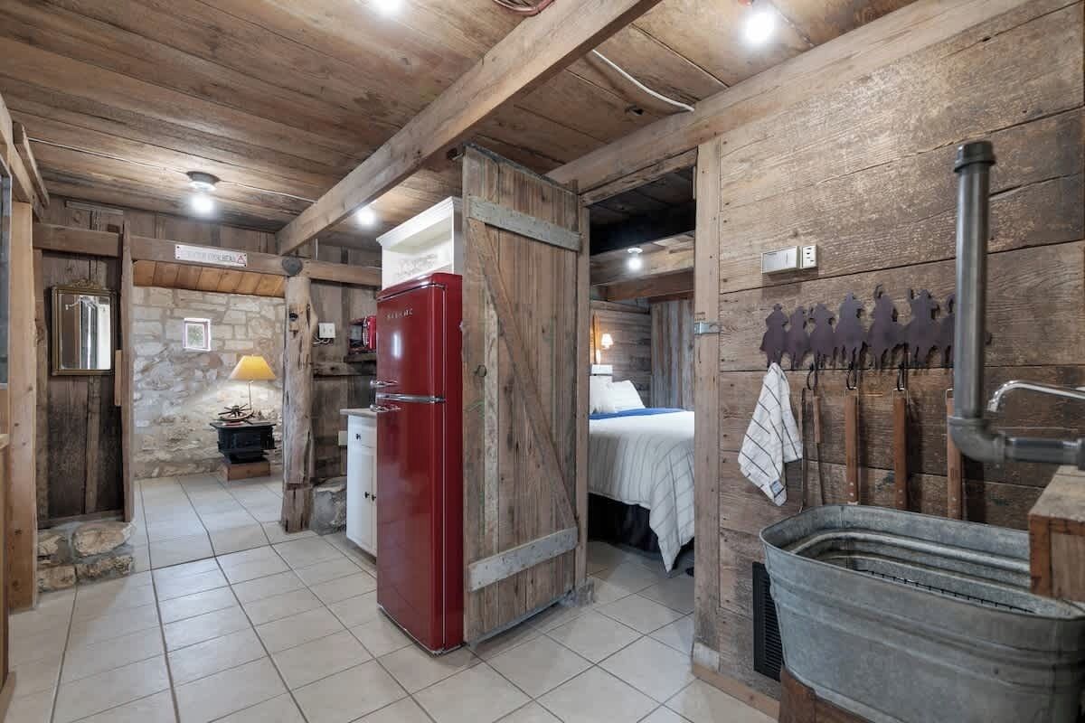 A rustic cabin interior with a stone wall, wooden beams, a red refrigerator, a bed, and a metal sink.