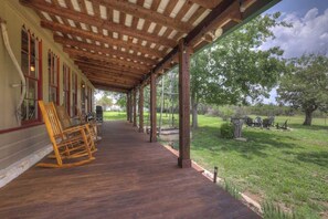 A covered porch with wooden flooring and rocking chairs, overlooking a grassy area with trees and outdoor furniture. - Private and Historic Rocking Ox Ranchette & Estate (Fredericksburg)