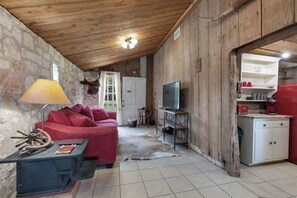 A cozy cabin interior with a stone wall, wooden ceiling, a red sofa, a television, and a small kitchen area. - Private and Historic Rocking Ox Ranchette & Estate (Fredericksburg)