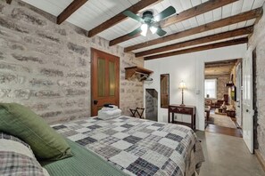 A bedroom with a stone wall, wooden beams, a bed with a quilt, a ceiling fan, and a view into another room. - Private and Historic Rocking Ox Ranchette & Estate (Fredericksburg)