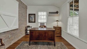 A room with a wooden desk, a chair, a small table, a lamp, and a window with a cross above it. - Private and Historic Rocking Ox Ranchette & Estate (Fredericksburg)