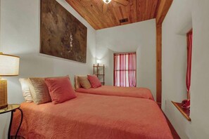 Una habitación con dos camas, un ventilador de techo, un cuadro abstracto grande, una ventana con cortinas rojas y una mesita con una lámpara.