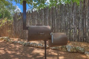 Un barbecue en métal doté d’une cheminée verticale, placé contre une clôture en bois et entouré de rochers.