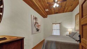 Ein Schlafzimmer mit Holzboden, einem Bett mit grauer Decke, einem Schreibtisch mit Laptop und einem Bild an der Wand.