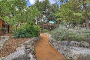 Un chemin de pierres mène à une construction en bois surmontée d’un toit rouge, le tout étant entouré d’une végétation luxuriante et d’arbres.