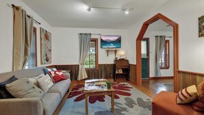 Ein Wohnzimmer mit einer Couch, einem Couchtisch und einem Bücherregal. An der Wand hängt ein Gemälde und ein Fenster ist mit Vorhängen versehen.