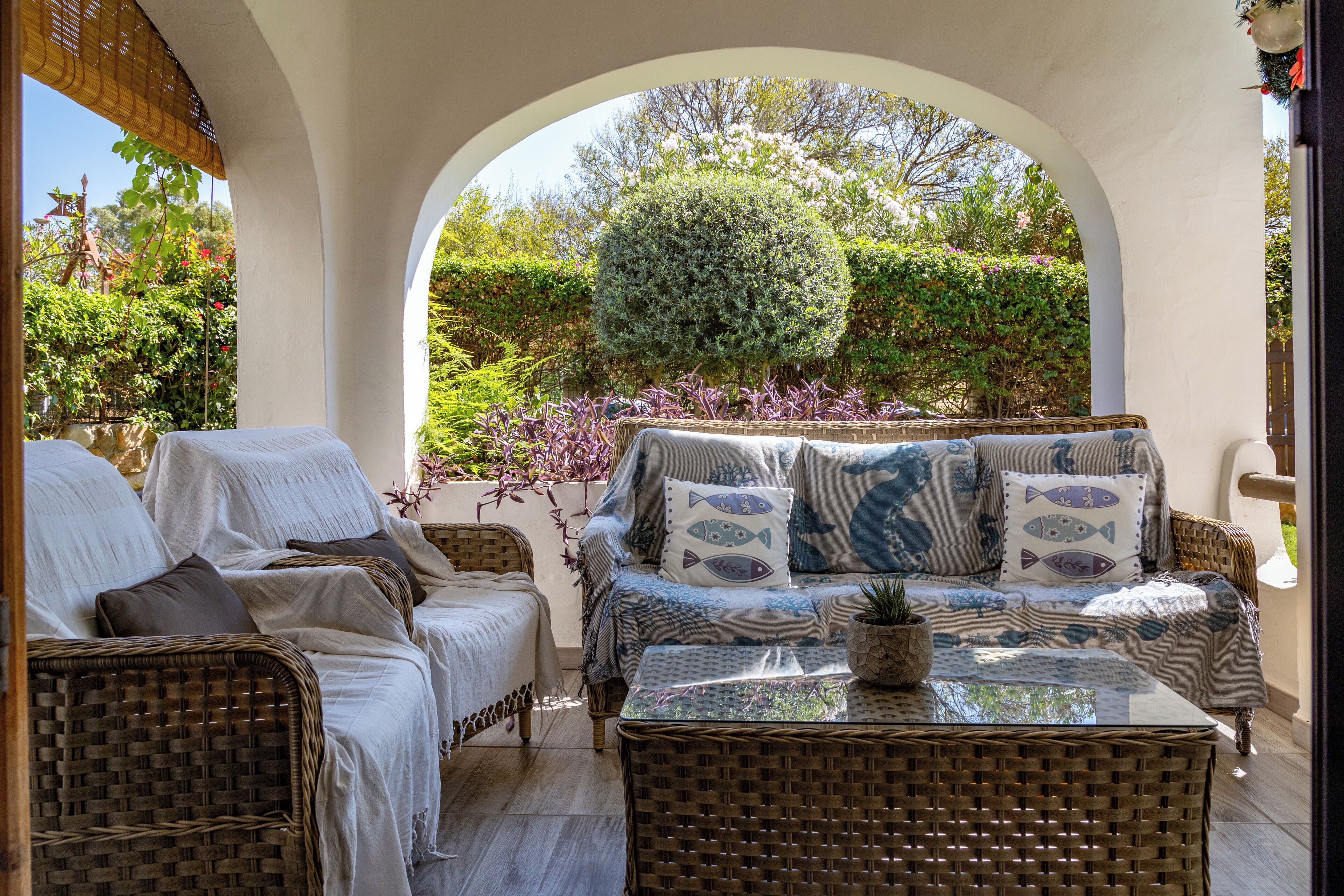 A sunlit patio with wicker furniture, a small table with a plant, and a view of a garden through an archway.
