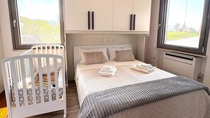 Et soveværelse med en vugge, en seng med et quiltet sengetæppe og et stort vindue med udsigt over grønne omgivelser.