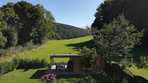 A rustic outdoor seating area with a wooden table and chairs, surrounded by lush greenery and a hill in the background.