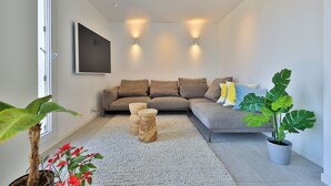 Ein modernes Wohnzimmer mit einem grauen Sofa, zwei Beistelltischen, einer großen Kühlpflanze und einem flachbildfernseher, der an der Wand montiert ist.