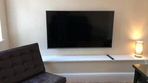 Una sala de estar con una televisión de pantalla plana montada en la pared, un sofá con cojines y una mesita de noche con una lámpara.