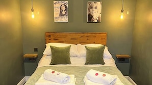 Quarto de hotel com cabeceira de madeira, duas almofadas verdes, cama com lençóis brancos, duas toalhas enroladas com botões vermelhos e duas luminárias fixadas na parede.