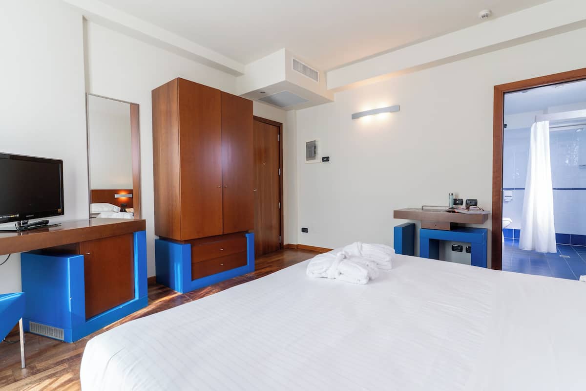 A hotel room with a bed, a TV, a wooden wardrobe, a desk with a computer, and a bathroom visible through an open door.