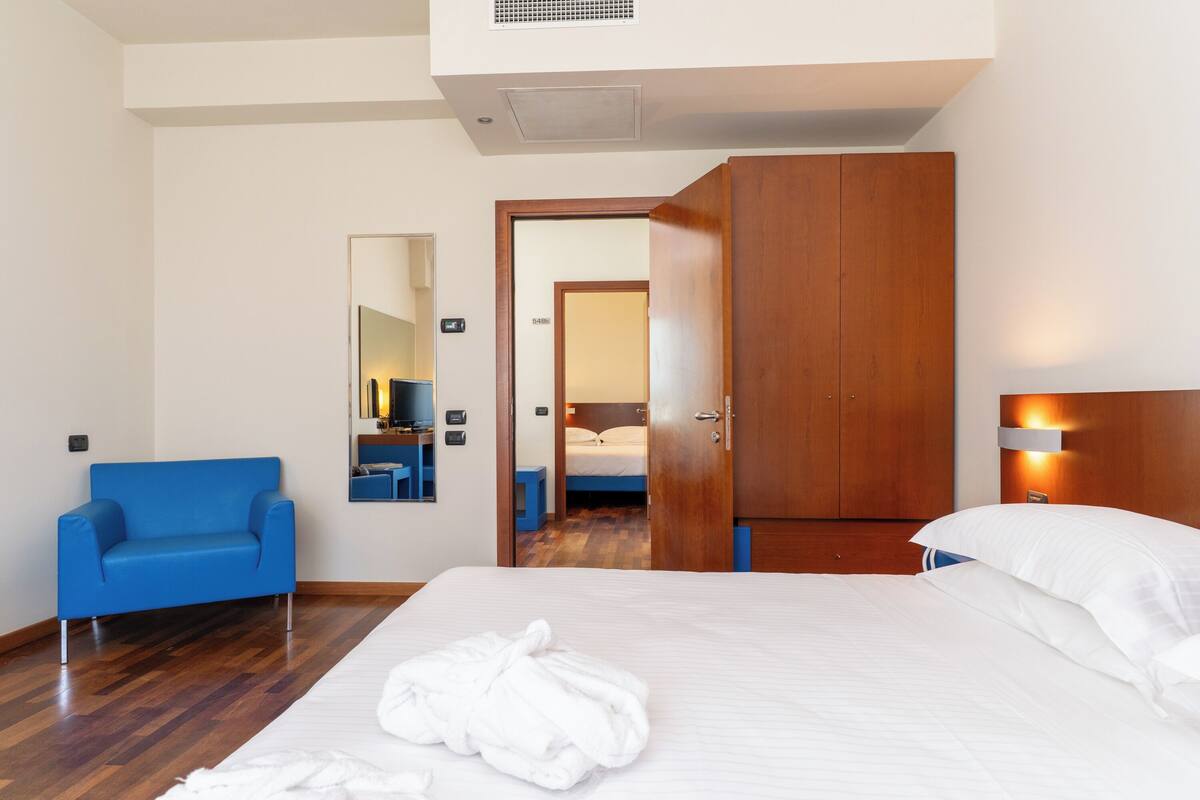 A hotel room with a bed, a blue chair, a wooden wardrobe, a mirror, and a wall-mounted light fixture.