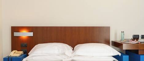 Une chambre d’hôtel avec une tête de lit en bois, une table de chevet avec un téléphone, une lampe et une petite étagère sur laquelle se trouvent une bouteille d’eau et des lunettes de soleil.