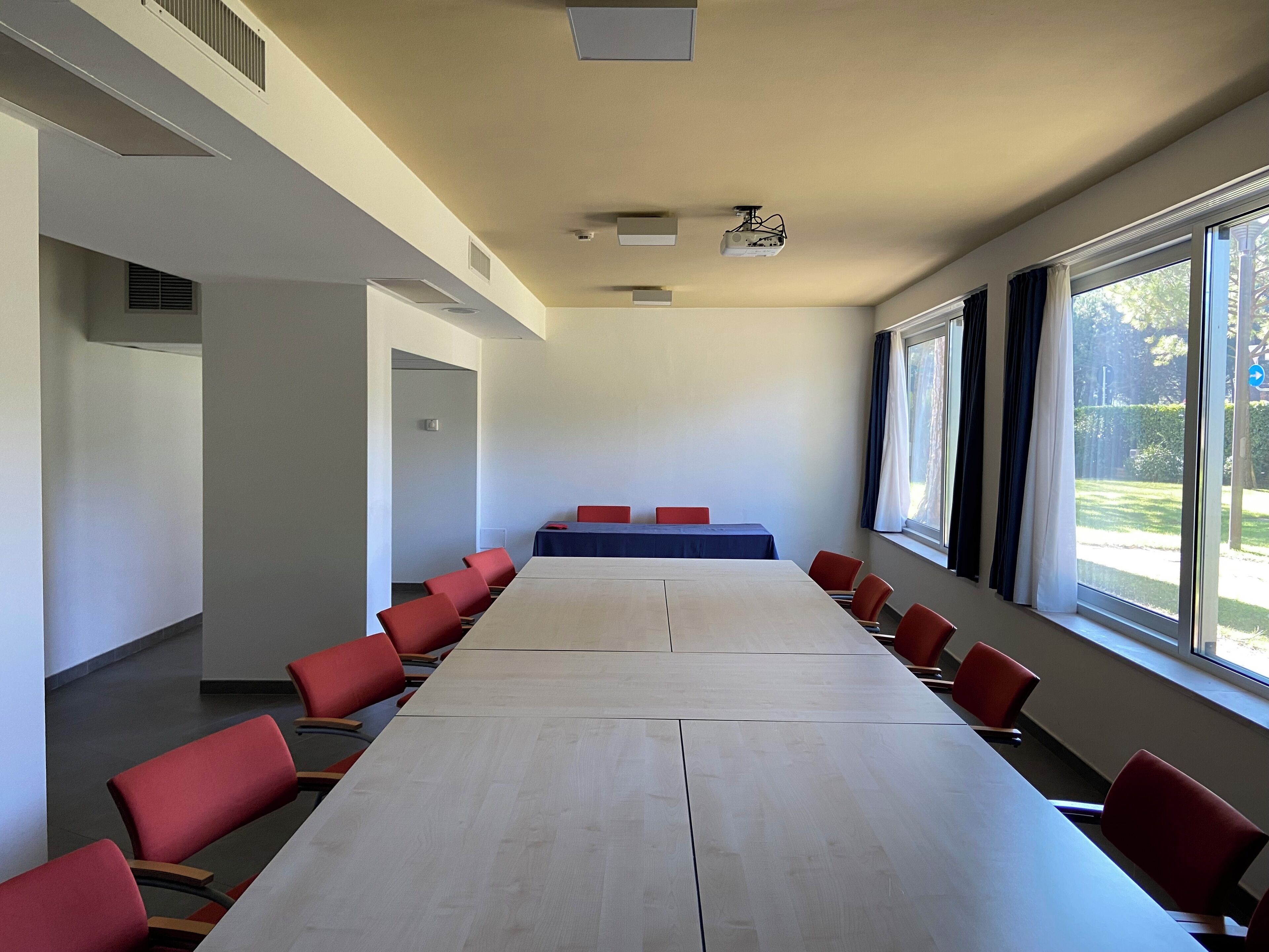 Een vergaderzaal met een lange tafel en rode stoelen, grote ramen en een projecteerder aan het plafond.