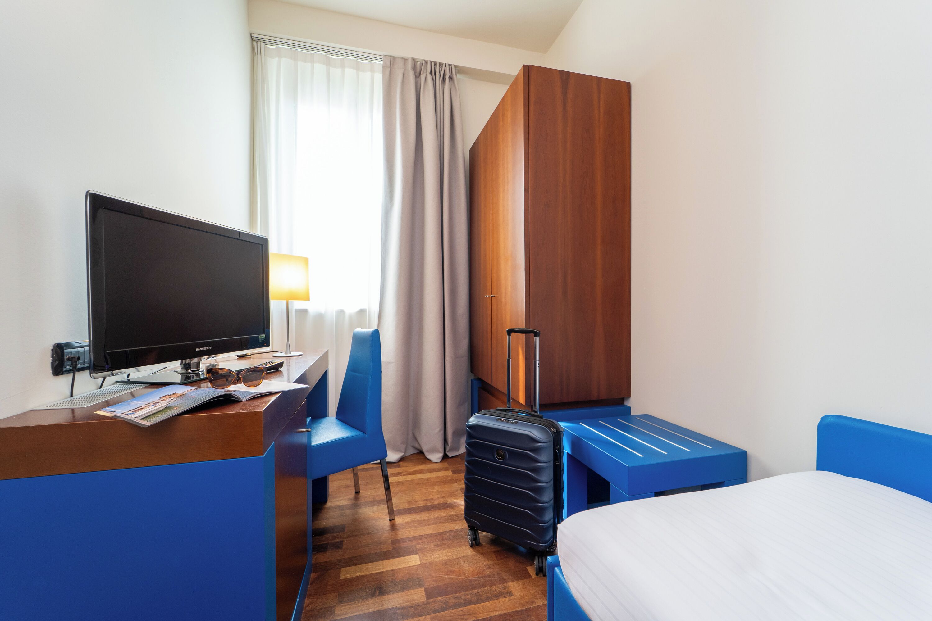 Una habitación de hotel con escritorio, televisión, cama, maleta y armario.