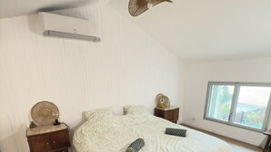 Una camera da letto con un letto grande, pavimento in legno, pareti bianche, una ventola a soffitto e una finestra con vista.