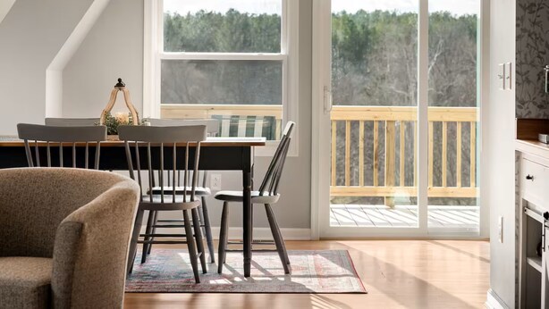 A dining area with a table, chairs, and a view of a wooden deck through large windows. - MountainWaters - Mtn, River, Relax! - Hottub, Private Riverfront, Firepit. (Maurertown)