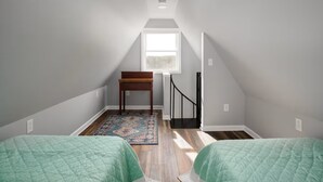 A small, well-lit attic bedroom with a sloped ceiling, a window, a wooden desk, a black railing, and two beds with green bedding. - MountainWaters - Mtn, River, Relax! - Hottub, Private Riverfront, Firepit. (Maurertown)